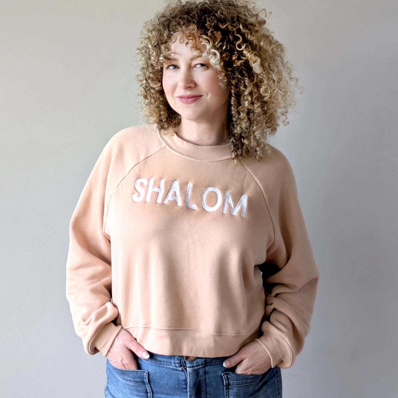 A person wearing a beige sweatshirt with the word 'SHALOM' written across the front in white letters, paired with blue jeans.
